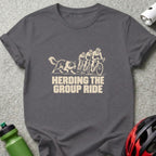 A dark gray crewneck t-shirt featuring a graphic of three cyclists and a herding dog, with the text "HERDING THE GROUP RIDE."