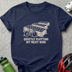A dark blue t-shirt printed with a bicycle, raised garden beds, and text reading QUIETLY PLOTTING MY NEXT RIDE.