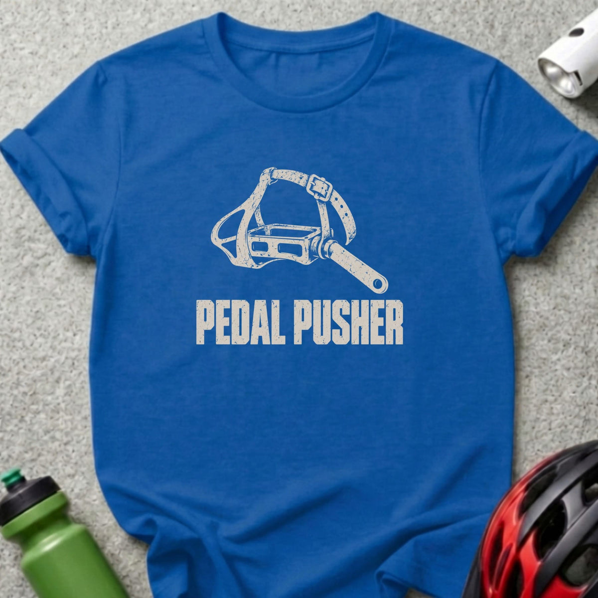 A blue t-shirt featuring a distressed white graphic of a bicycle pedal with a toe strap and the text PEDAL PUSHER.