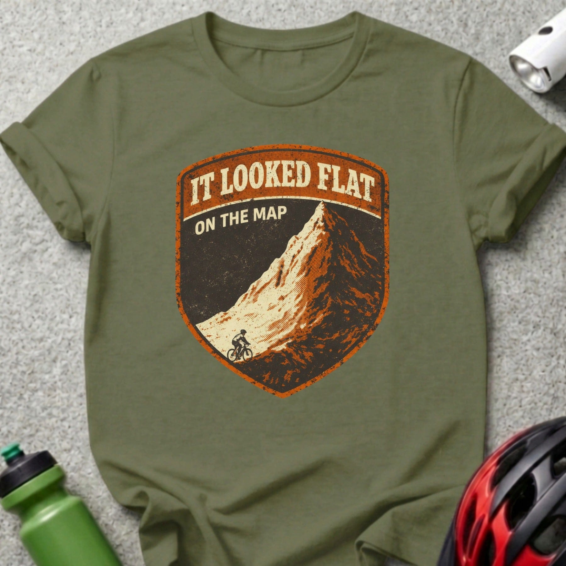 Olive green t-shirt with distressed shield logo featuring mountain and cyclist, text "IT LOOKED FLAT ON THE MAP".
