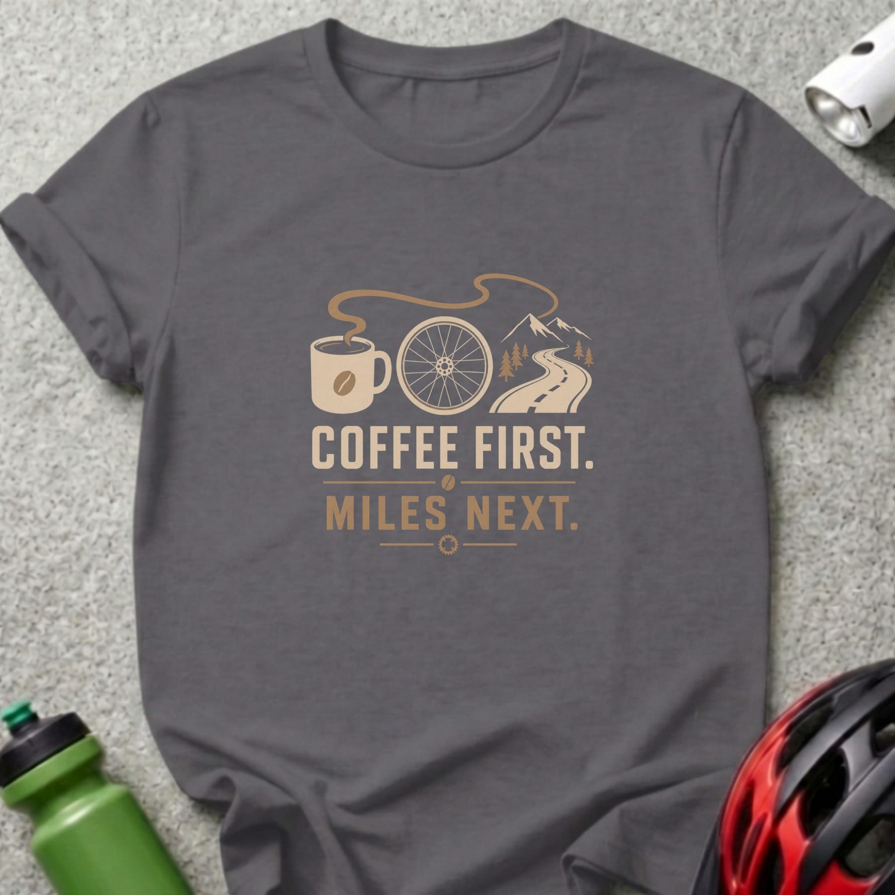 T-shirt with graphic of coffee cup, bike wheel, road, and mountains. Text reads "Coffee First. Miles Next."