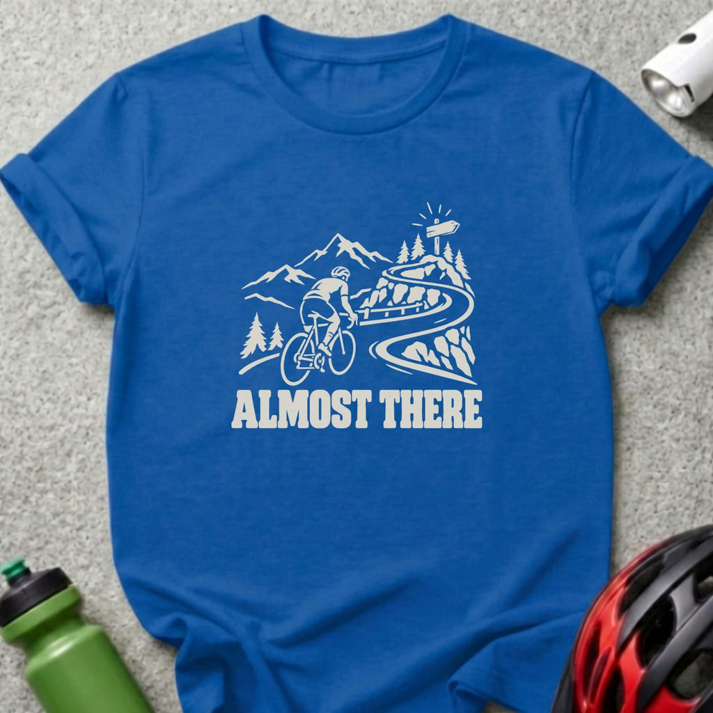 A blue t-shirt featuring a white graphic of a cyclist climbing a mountain road with the text ALMOST THERE.