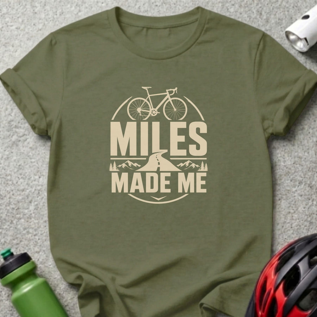 Olive green t-shirt with a beige graphic of a bicycle, road, and mountains reading "MILES MADE ME".