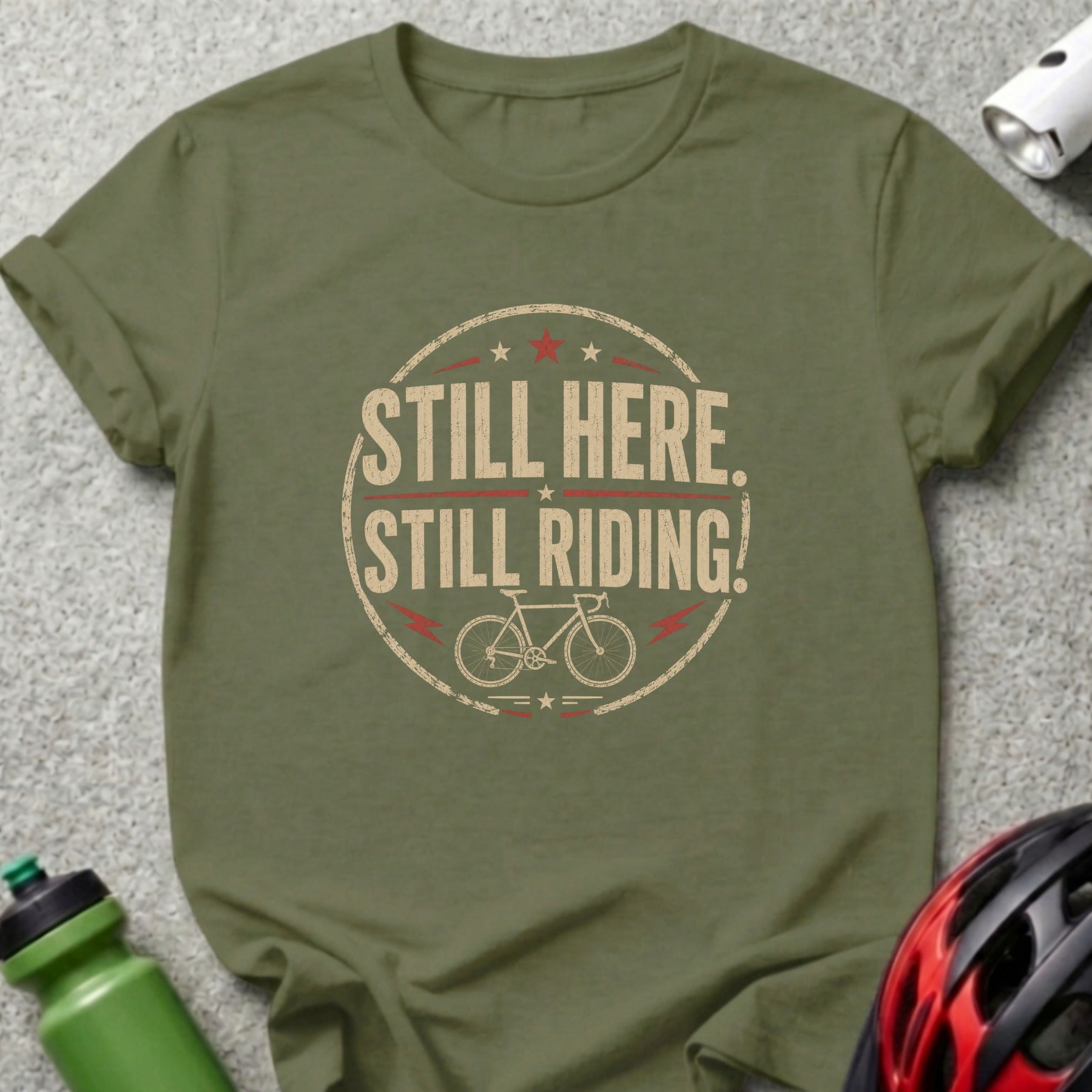 Olive green t-shirt with "STILL HERE. STILL RIDING!" text and a bicycle graphic.