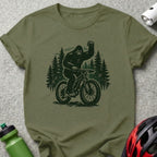 Olive t-shirt featuring a Bigfoot riding a mountain bike with trees behind it.