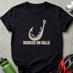 A black t-shirt features a cream graphic of a fishhook shaped like a winding mountain road with a cyclist and the text "HOOKED ON HILLS".