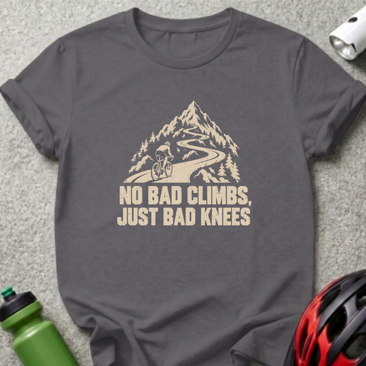 A charcoal t-shirt featuring a graphic of a cyclist climbing a winding mountain road with the text "NO BAD CLIMBS, JUST BAD KNEES."