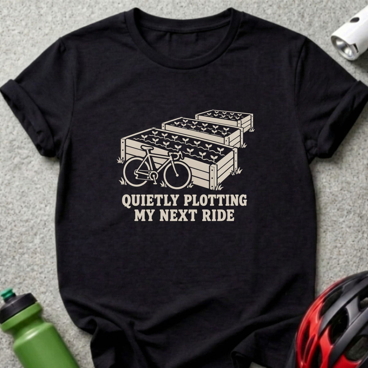 A black t-shirt printed with raised garden beds, a bicycle, and the text "QUIETLY PLOTTING MY NEXT RIDE".