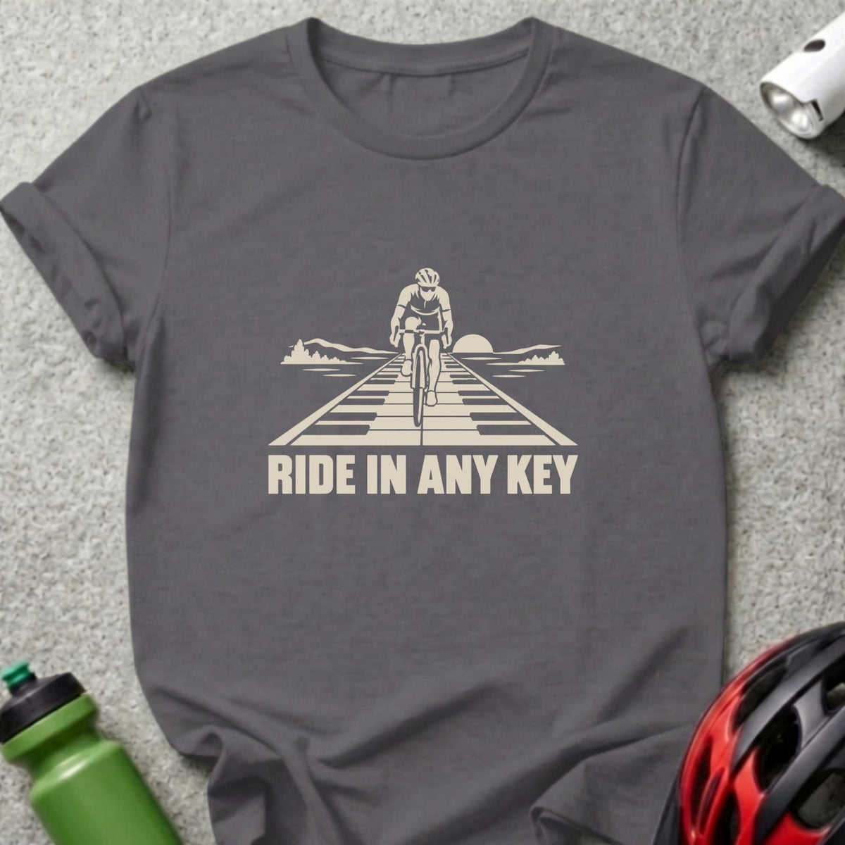 Charcoal grey t-shirt featuring a graphic of a cyclist riding on a piano keyboard road with text reading RIDE IN ANY KEY.