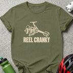 An olive green t-shirt featuring a beige graphic of a fishing reel with a bicycle pedal handle and the text REEL CRANKY.