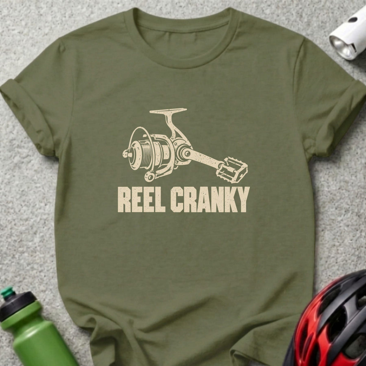 An olive green t-shirt featuring a beige graphic of a fishing reel with a bicycle pedal handle and the text REEL CRANKY.