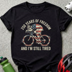 Black t-shirt with cyclist and American flag graphic, text reads "250 Years of Freedom and I'm Still Tired."