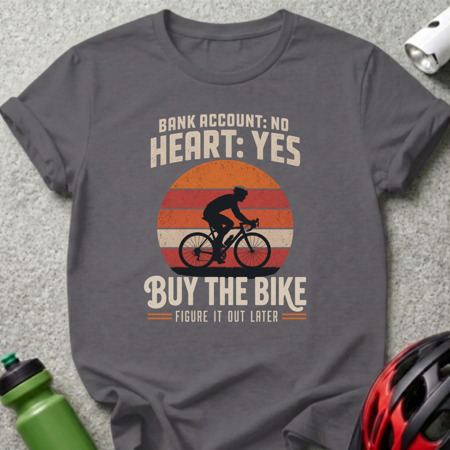 Dark gray t-shirt with retro sunset cyclist graphic and text: "Bank account: No. Heart: Yes. Buy the bike. Figure it out later."