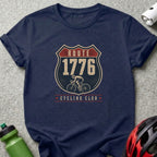 Navy t-shirt with "Route 1776 Cycling Club" and cyclist graphic on a shield.