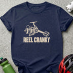 Dark blue t-shirt featuring a graphic of a fishing reel with a bicycle pedal handle and the text REEL CRANKY.