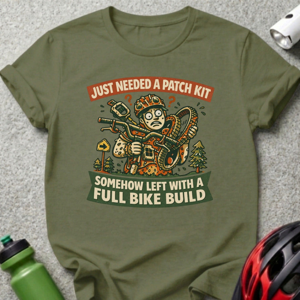 T-shirt graphic with cartoon cyclist holding flat tire, text: "Just Needed a Patch Kit, Somehow Left with a Full Bike Build".