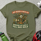 T-shirt graphic with cartoon cyclist holding flat tire, text: "Just Needed a Patch Kit, Somehow Left with a Full Bike Build".