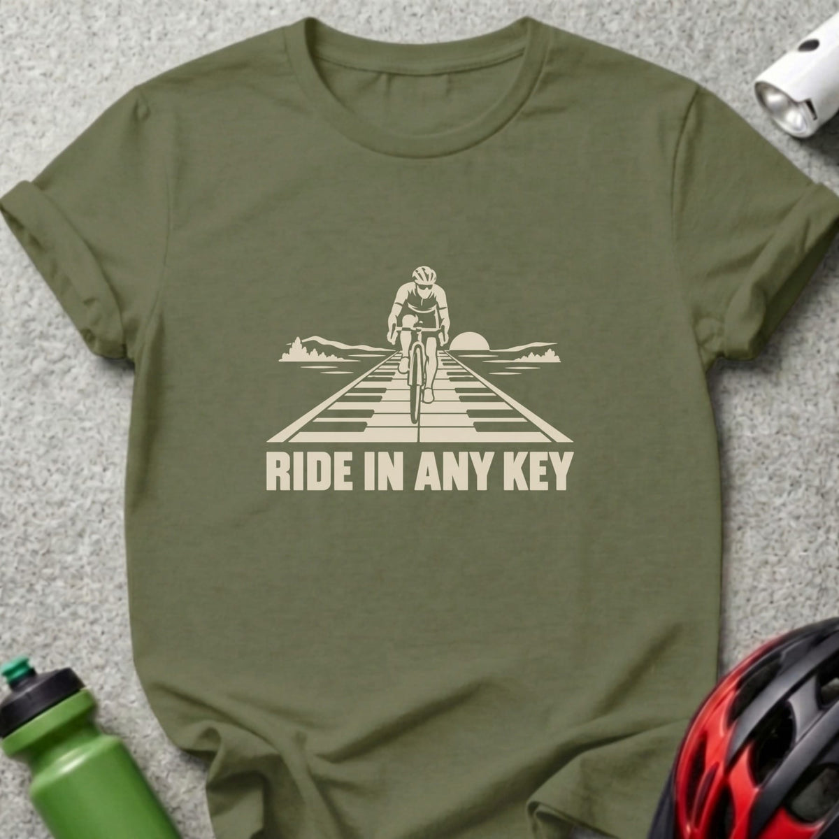 An olive green t-shirt featuring a graphic of a cyclist riding on piano keys and the text RIDE IN ANY KEY.