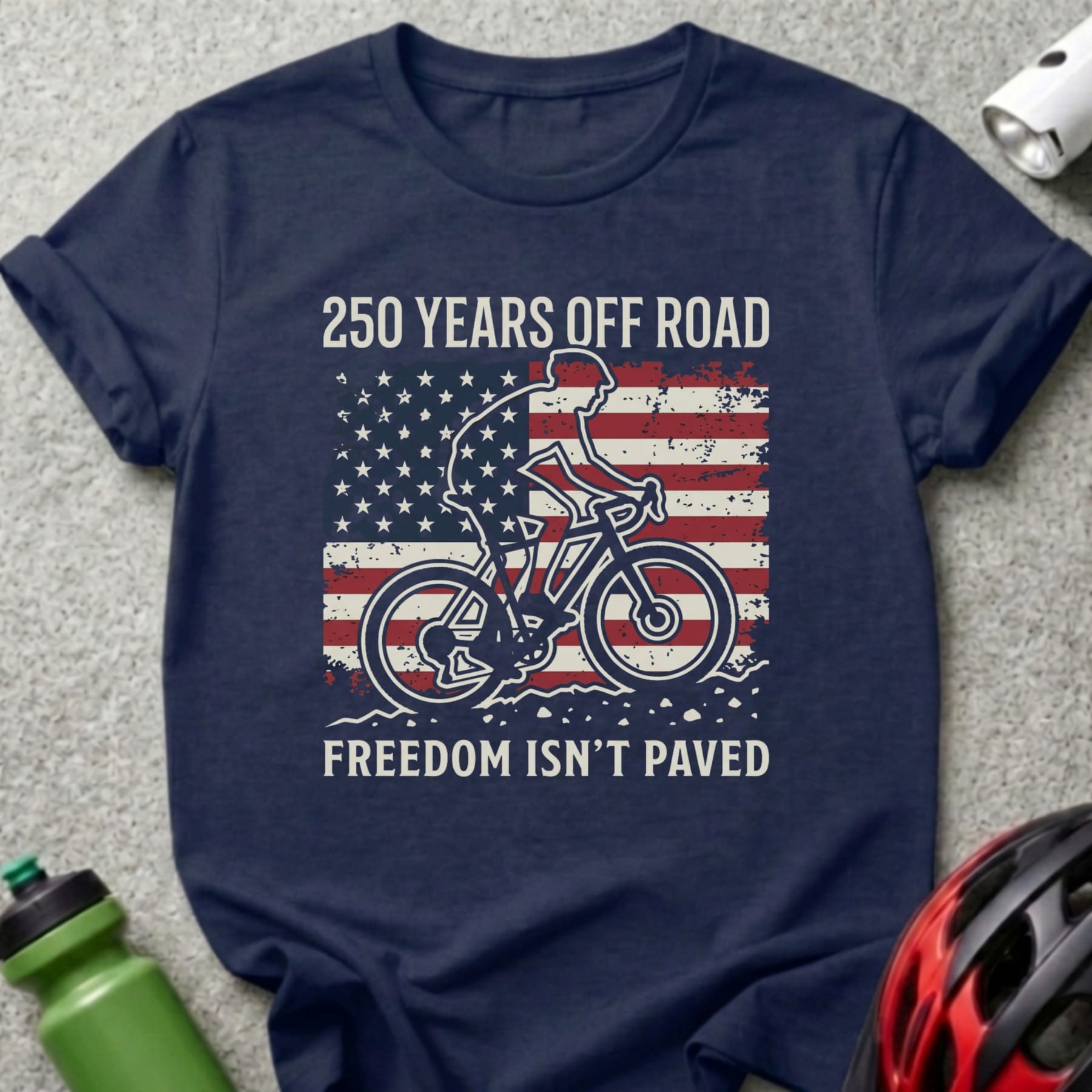 Navy t-shirt with cyclist silhouette on American flag, reads "250 Years Off Road Freedom Isn't Paved".