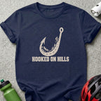 A navy blue t-shirt printed with a fishhook-shaped winding road graphic and the text "HOOKED ON HILLS".