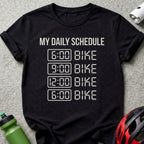 Black t-shirt with white text "MY DAILY SCHEDULE" and "6:00 BIKE", "9:00 BIKE", "12:00 BIKE", "6:00 BIKE".
