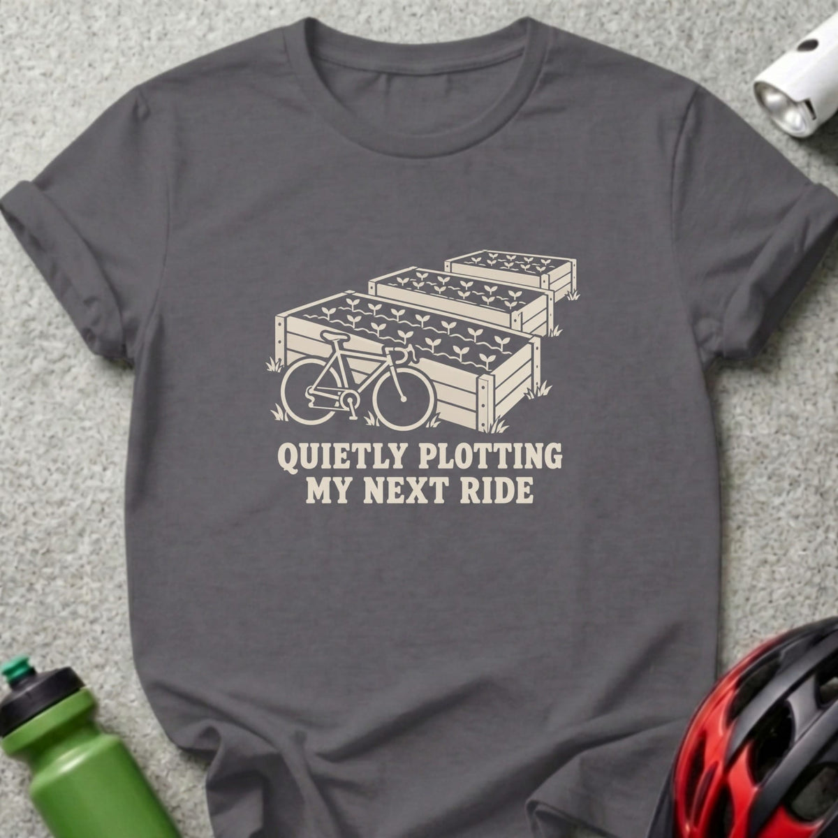 Dark gray t-shirt featuring a graphic of raised garden beds, a bicycle, and the text QUIETLY PLOTTING MY NEXT RIDE.