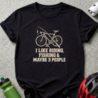 A black t-shirt featuring a bicycle and fishing rod graphic above the text, I LIKE RIDING, FISHING & MAYBE 3 PEOPLE.
