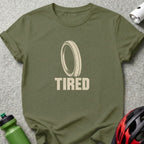 An olive green t-shirt with a bicycle tire graphic and the text TIRED underneath.