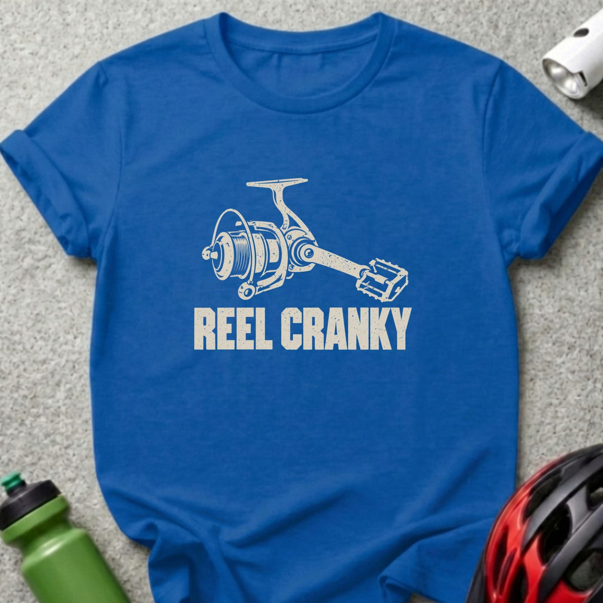 A royal blue t-shirt featuring a white graphic of a fishing reel with a bicycle pedal handle and the text REEL CRANKY.