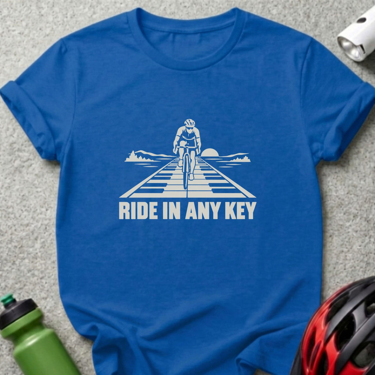 A blue t-shirt featuring a white graphic of a cyclist on piano keys and the text RIDE IN ANY KEY.