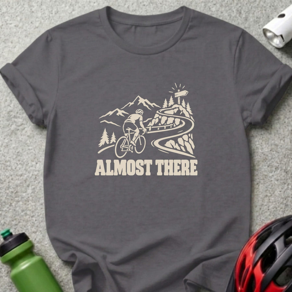 Charcoal gray t-shirt featuring a white graphic of a cyclist climbing a mountain road with the text ALMOST THERE.