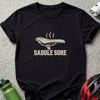 A black t-shirt featuring a beige graphic of a bicycle saddle with steam rising from it, above the text SADDLE SORE.