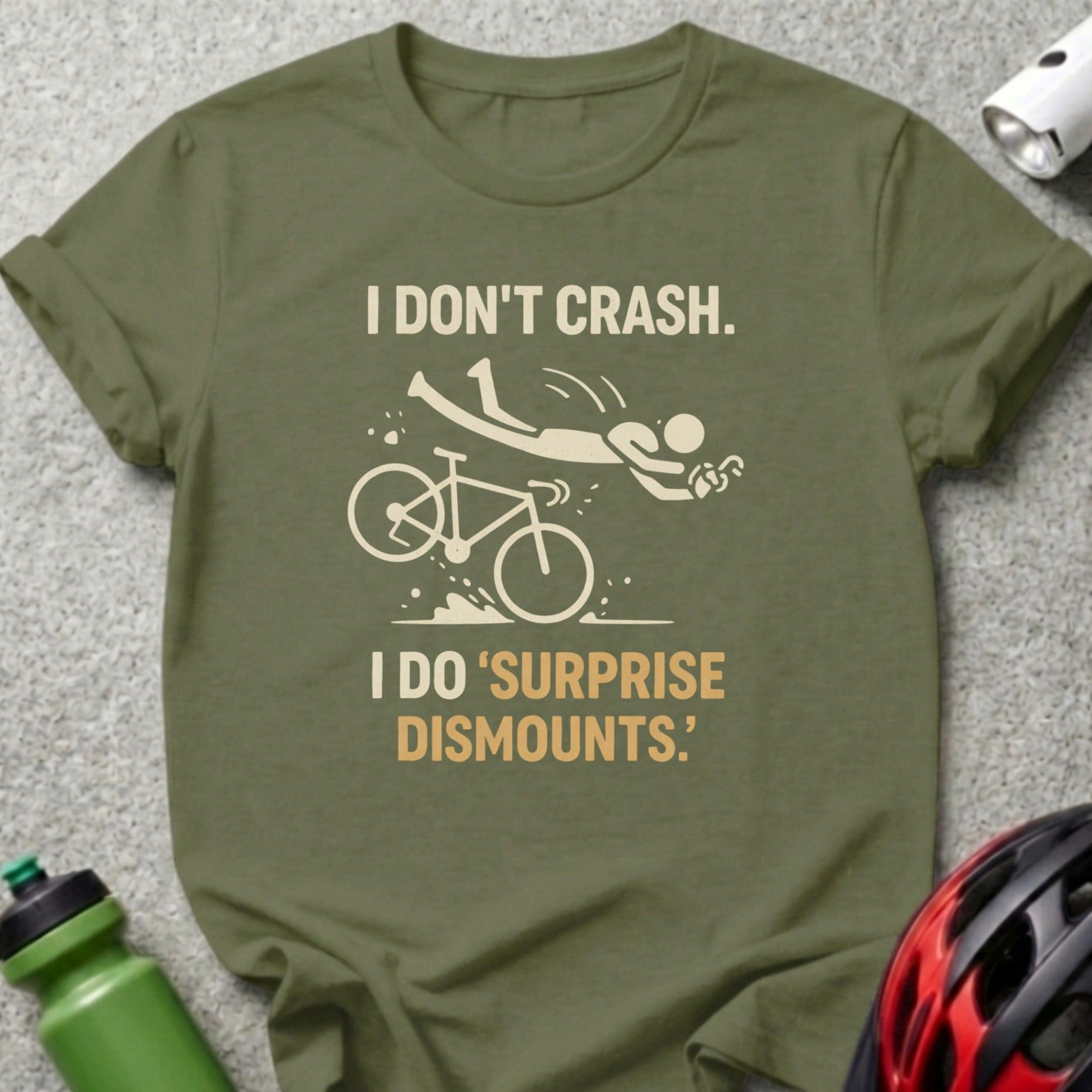 Olive green t-shirt with text "I DON'T CRASH. I DO 'SURPRISE DISMOUNTS.'" and a cyclist illustration.